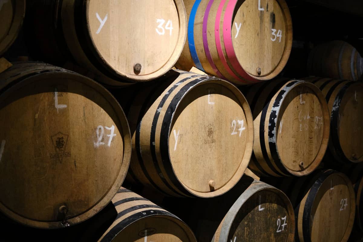 Uma pilha de barris de madeira com marcações de giz armazenados em um escuro porão de cerveja lambic, alguns com faixas coloridas, evocando a tradição de fermentação espontânea.
