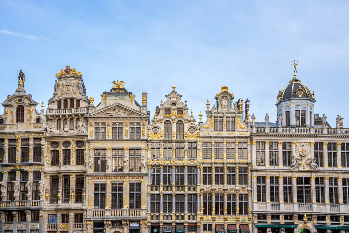 Fileira de ornamentadas guildas históricas com detalhes dourados na Grand Place de Bruxelas sob um céu azul parcialmente nublado.