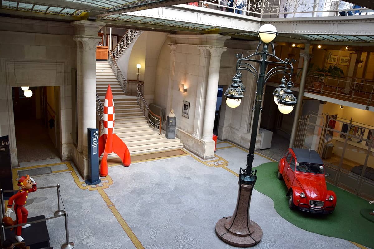 Vista do átrio central do Centro Belga de Histórias em Quadrinhos, com arquitetura Art Nouveau, um modelo de foguete vermelho, um carro antigo e estátuas de personagens de quadrinhos.