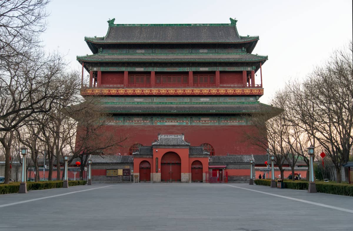 Der Trommelturm in Peking im warmen Nachmittagslicht, frontal aufgenommen mit seinen roten Mauern und dem grünen Dach, davor Bäume und ein offener Platz.