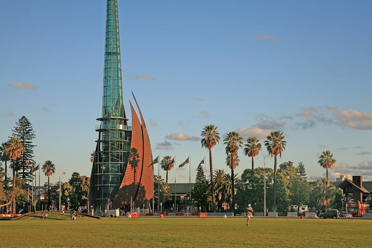 Weitwinkelbild des markanten Glas- und Kupferturms des Glockenturms (Swan Bells) an Perths Uferpromenade, umgeben von Palmen und einer Rasenfläche.