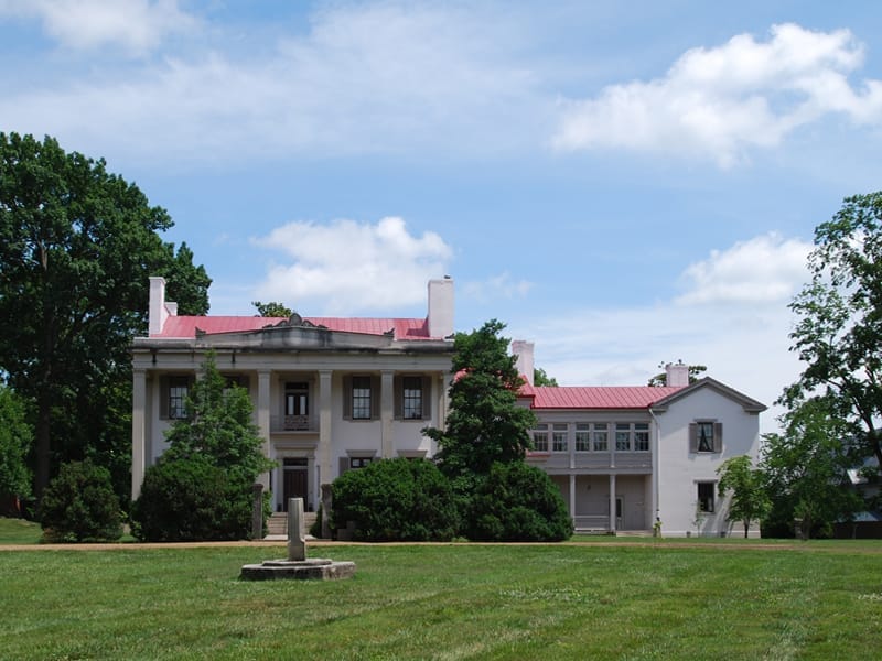 Mansión de Belle Meade Plantation con columnas blancas y techo rojo, rodeada de césped verde, arbustos y árboles altos en un día soleado.