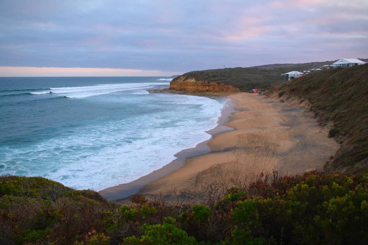 Vista panorámica de Bells Beach al atardecer con arena dorada, olas entrando, acantilados de arenisca y vegetación costera en primer plano, cerca de Torquay, Australia.