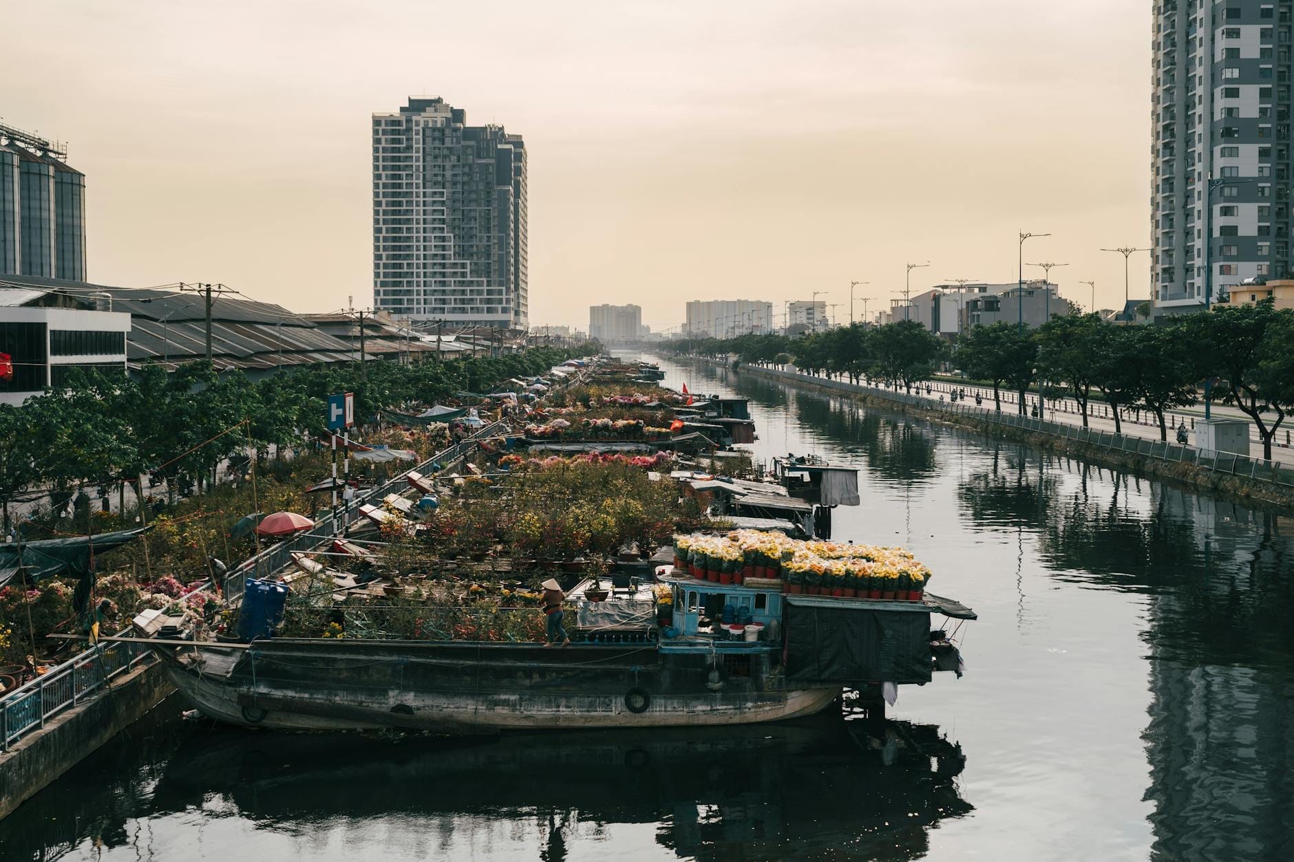 Un canal urbano tranquilo bordeado de barcas con flores y mercancías, flanqueado por edificios modernos y paseos arbolados al atardecer en Ciudad Ho Chi Minh.