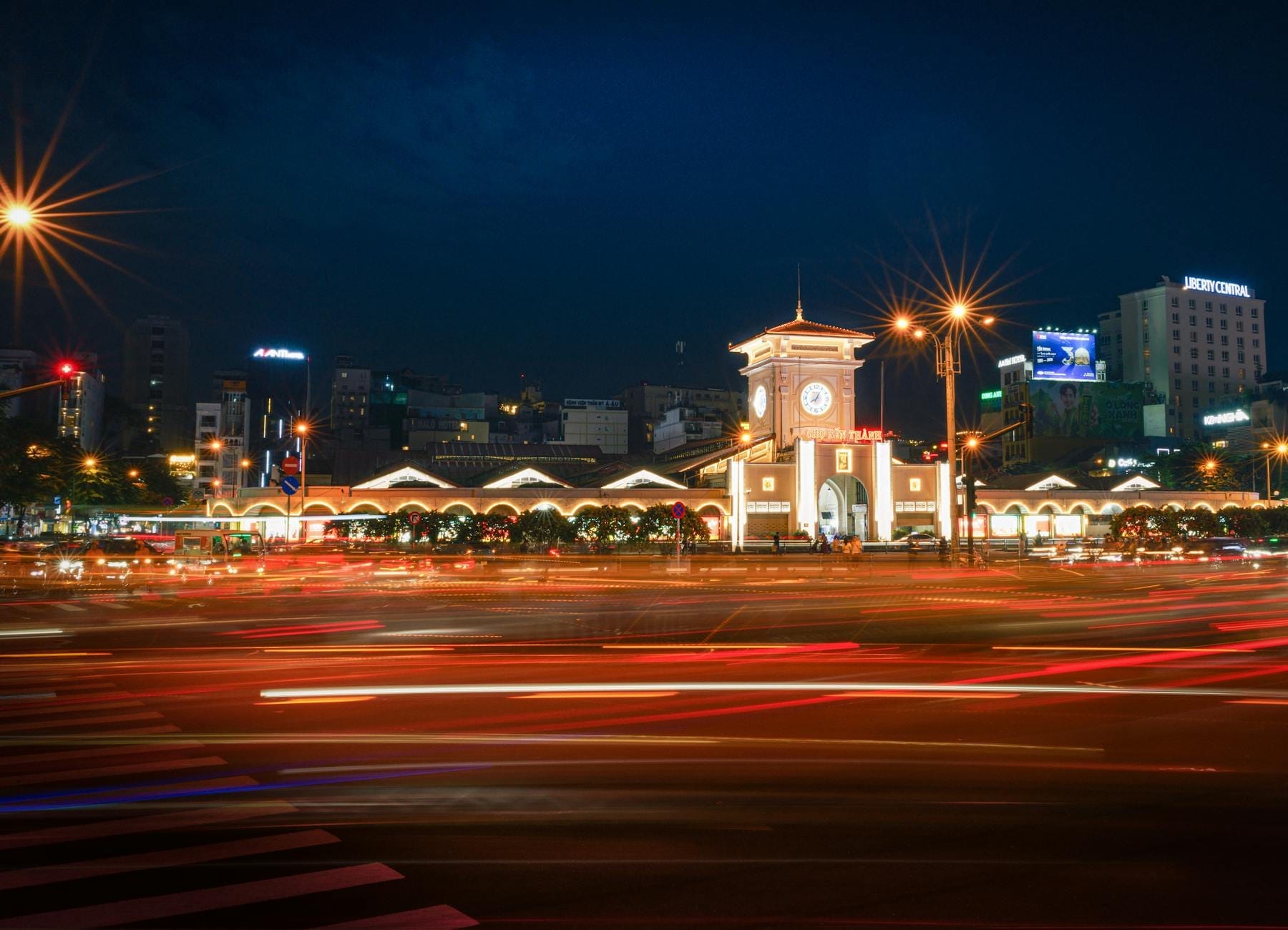 Vista nocturna del icónico mercado Bến Thành con su torre del reloj iluminada, una calle animada con rastros de luz y edificios de la ciudad al fondo.