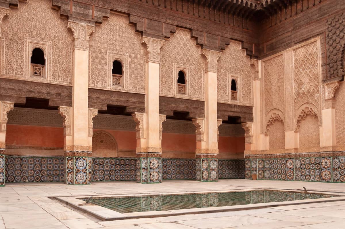 ลานกลางของ Ben Youssef Madrasa กับซุ้มประตูแกะสลัก กระเบื้องเซลลิจสีสันสดใส และสระน้ำสะท้อนรูปทรงสี่เหลี่ยมผืนผ้า