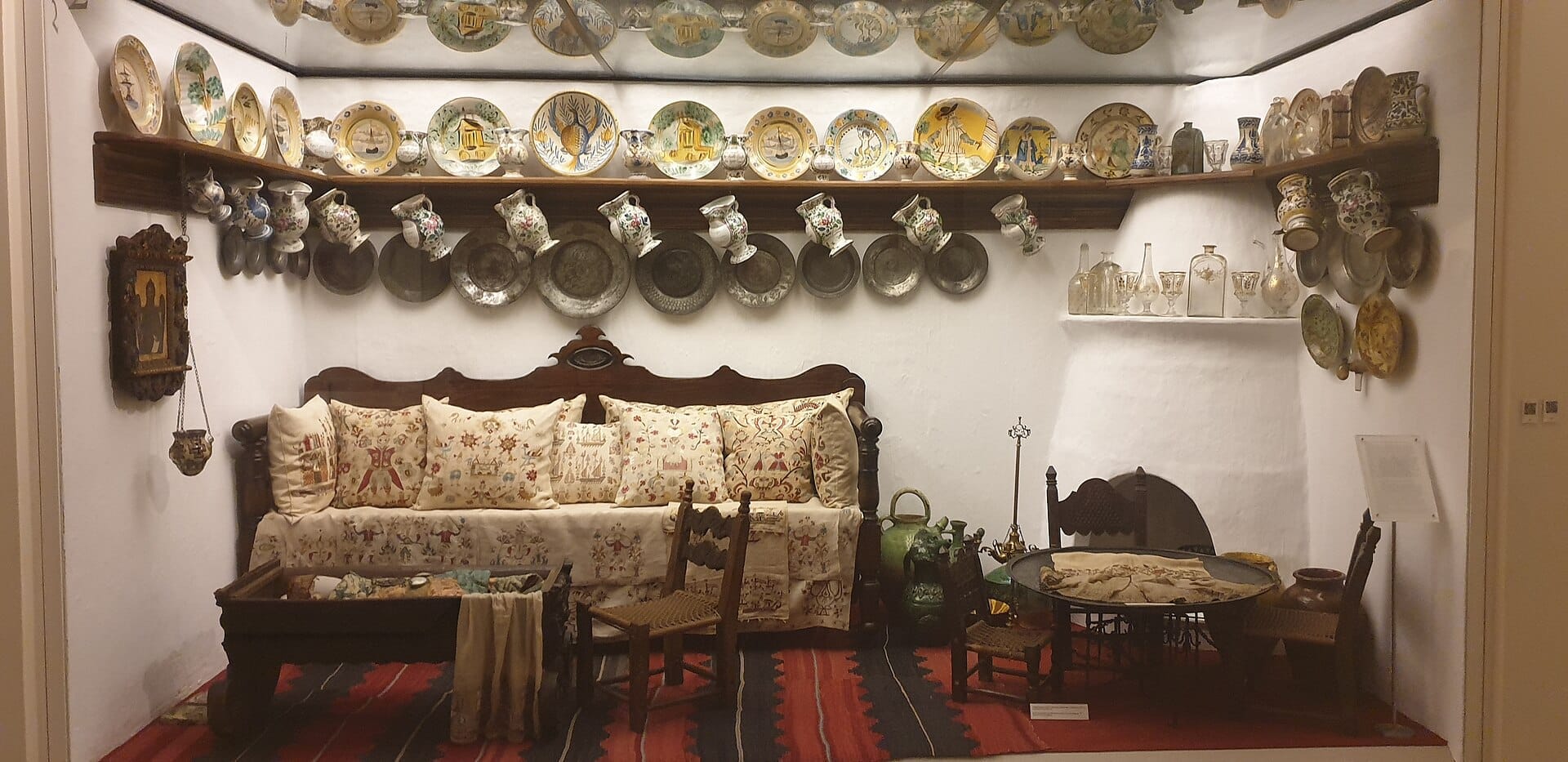 Sala greca tradizionale al Museo Benaki, con cuscini ricamati, mobili in legno e scaffali di ceramiche decorative su pareti imbiancate.