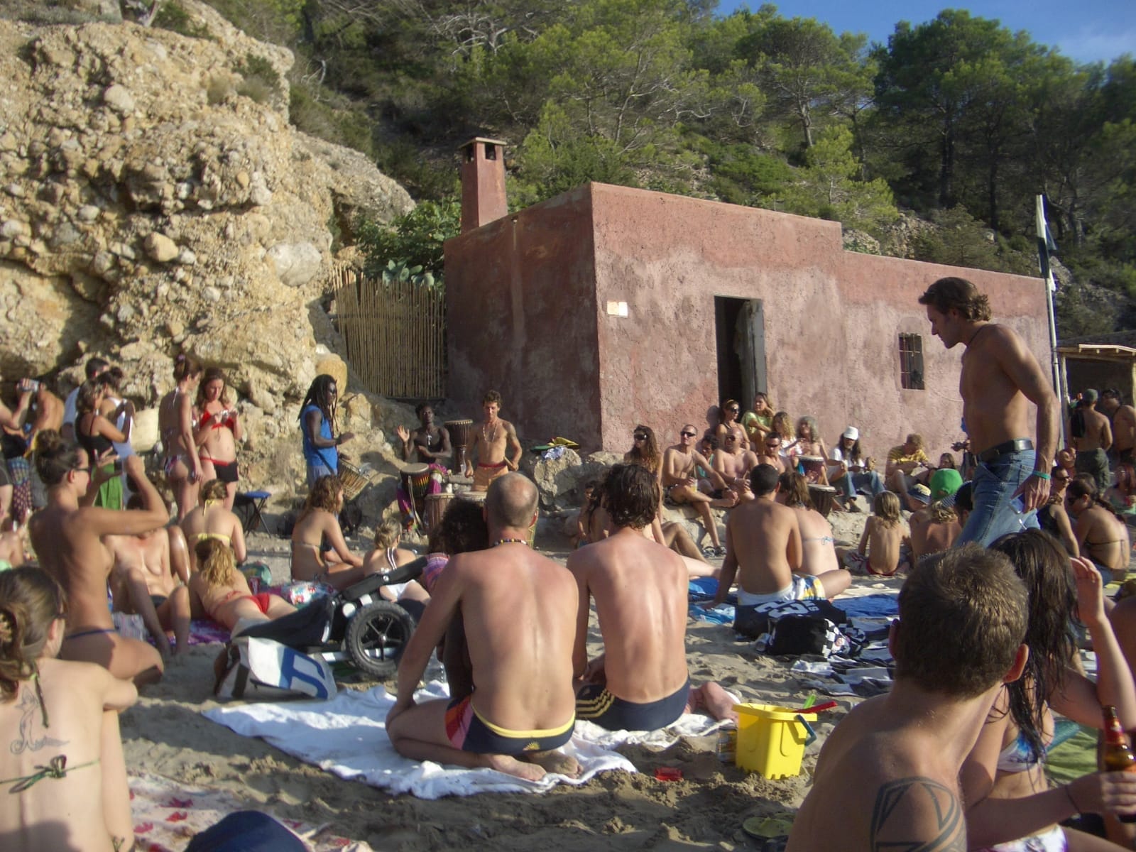 I visitatori si riuniscono sulla riva sabbiosa della spiaggia di Benirràs, Ibiza, rilassandosi vicino a un edificio rustico circondato da scogliere rocciose e pini.