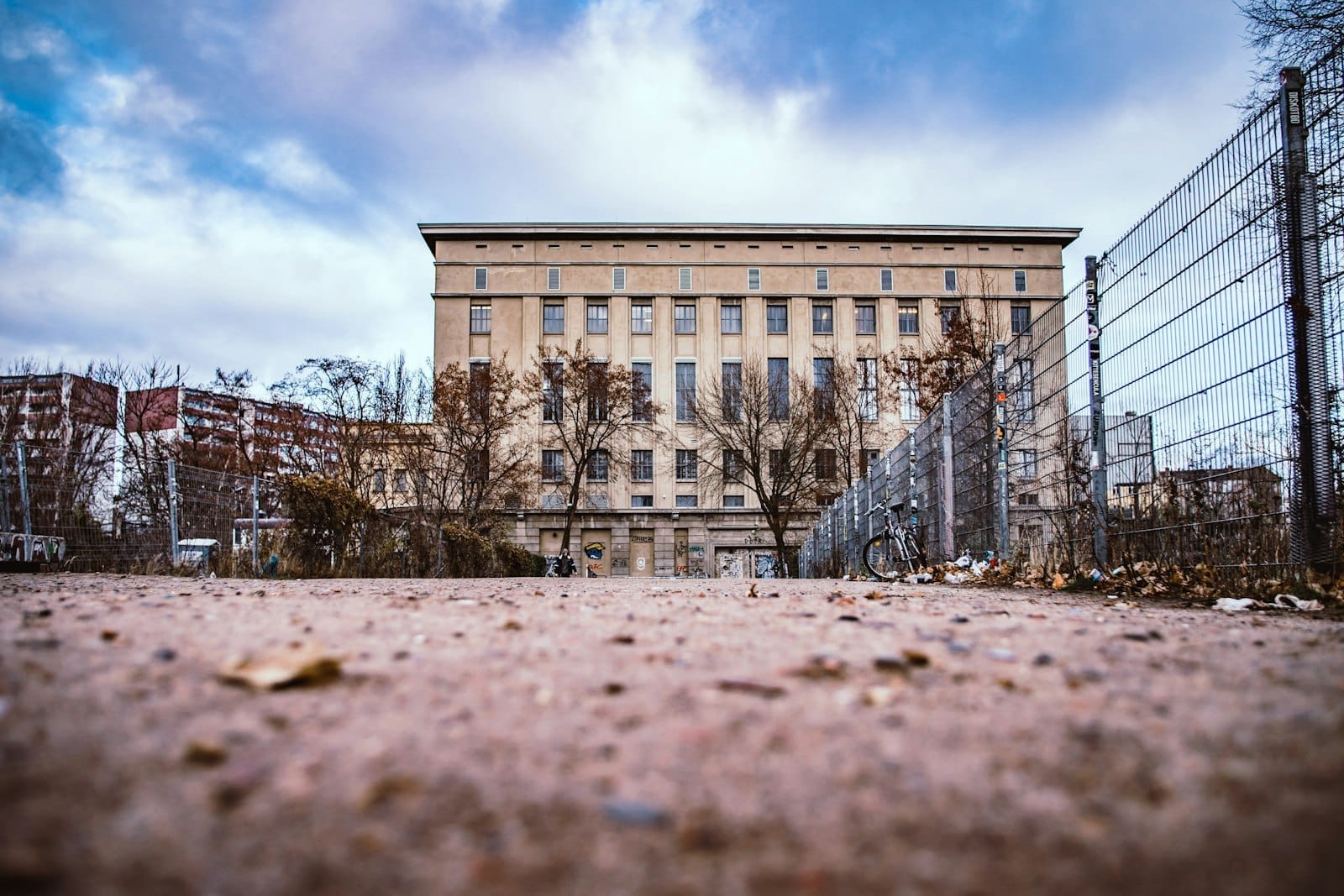 Vista ampia della facciata industriale del Berghain con cielo coperto, vialetto recintato, alberi spogli e una bicicletta all'ingresso.