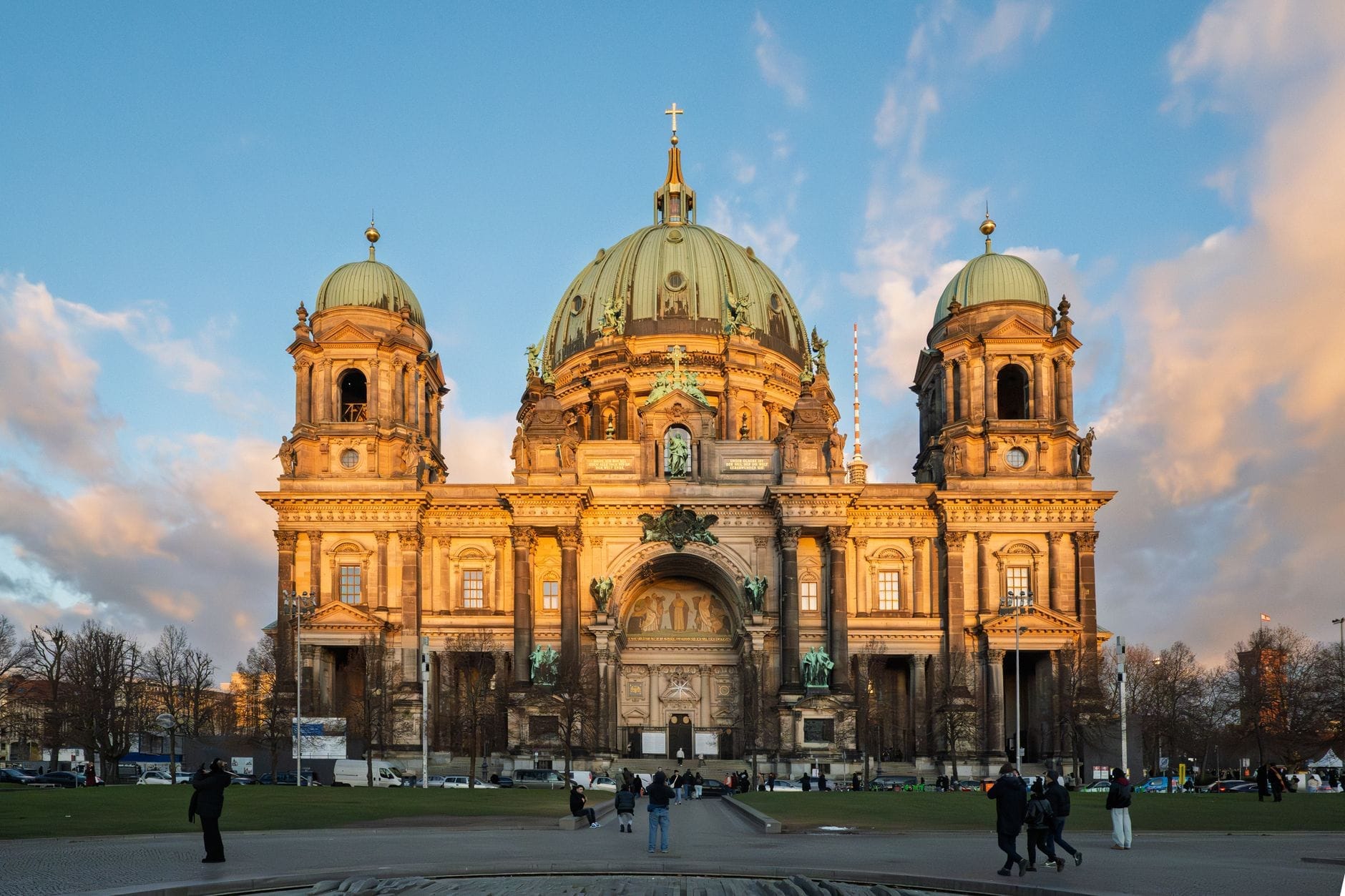 Vista frontale e ampia del Berliner Dom al tramonto, con la luce dorata sulla cupola e le torri e gruppi di persone che camminano e si radunano sul grande prato antistante.