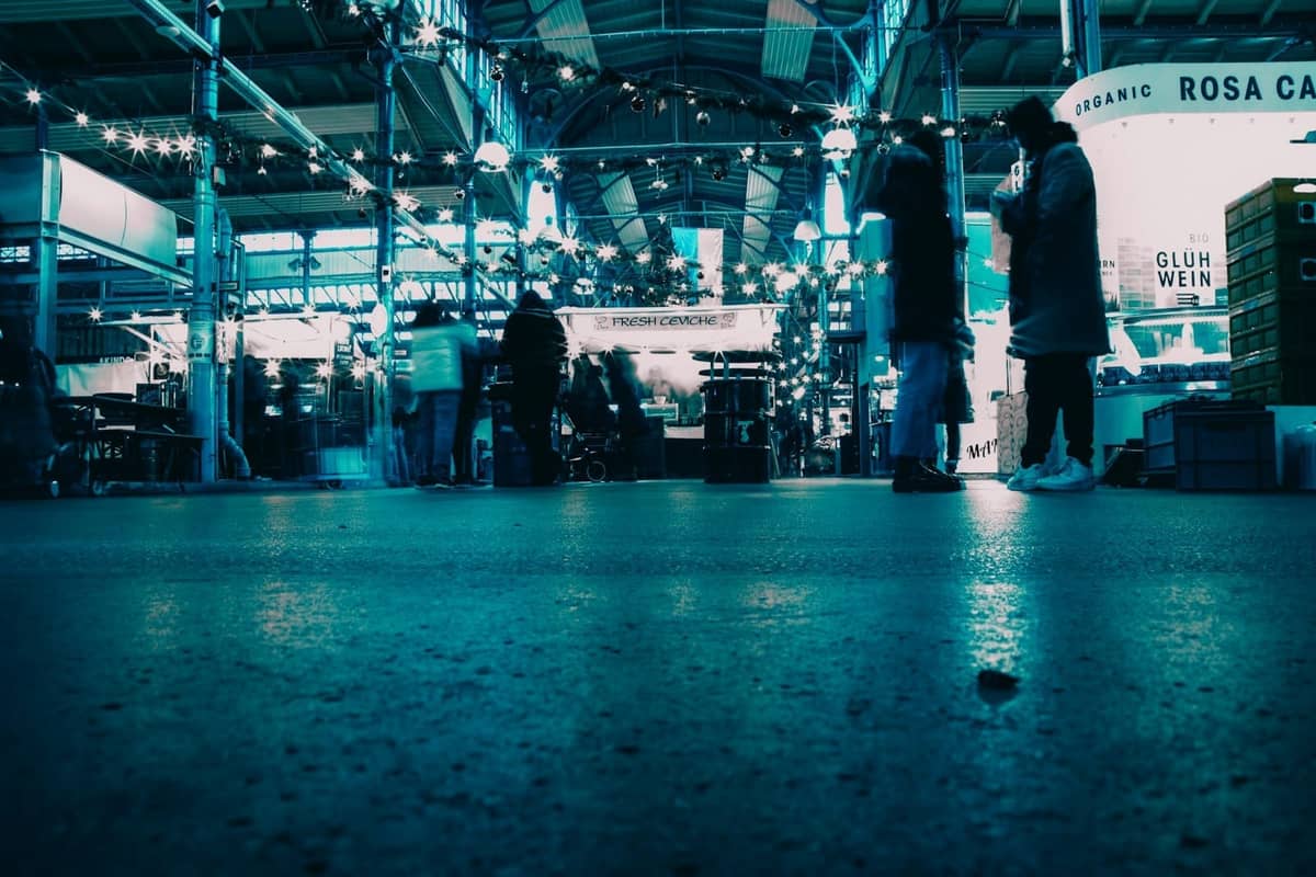 Szene in einem Berliner Innenmarkt mit Menschen, die sich unterhalten, beleuchteten Ständen und Lichterketten darüber, die eine lebhafte Atmosphäre der Essens- und Straßenkultur heraufbeschwören.