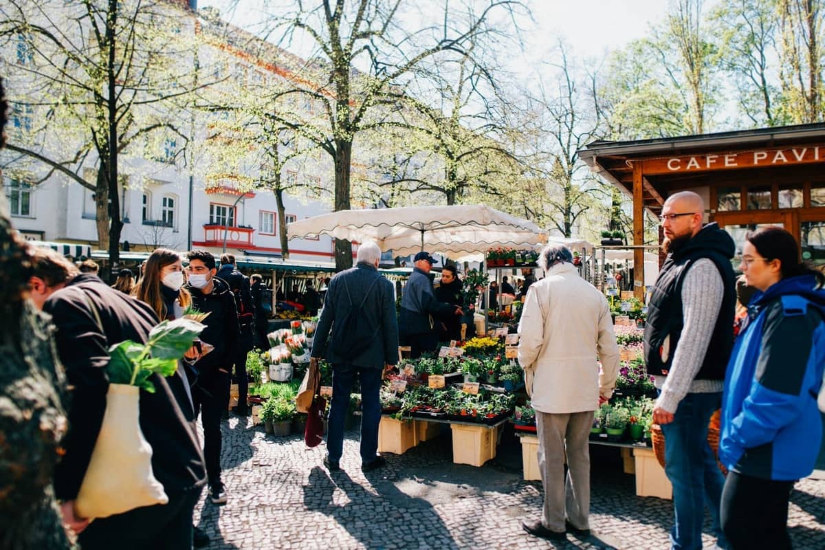 Menschen beim Einkaufen an Outdoor-Marktständen mit Blumen und Produkten, umgeben von Bäumen und Berliner Gebäuden an einem sonnigen Tag.