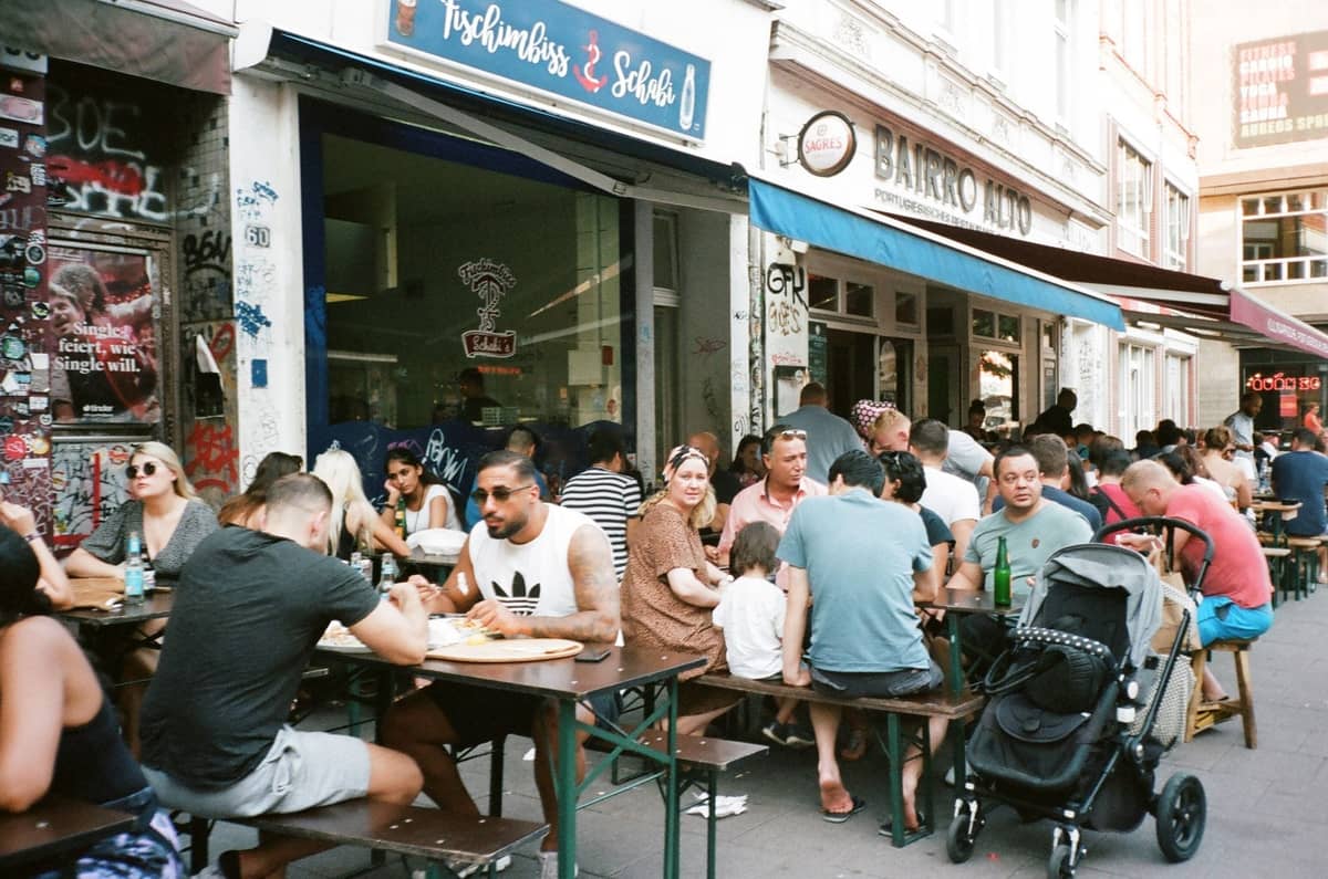 Animada escena de comedor al aire libre con personas comiendo en mesas fuera de restaurantes del barrio en una calle de Berlín