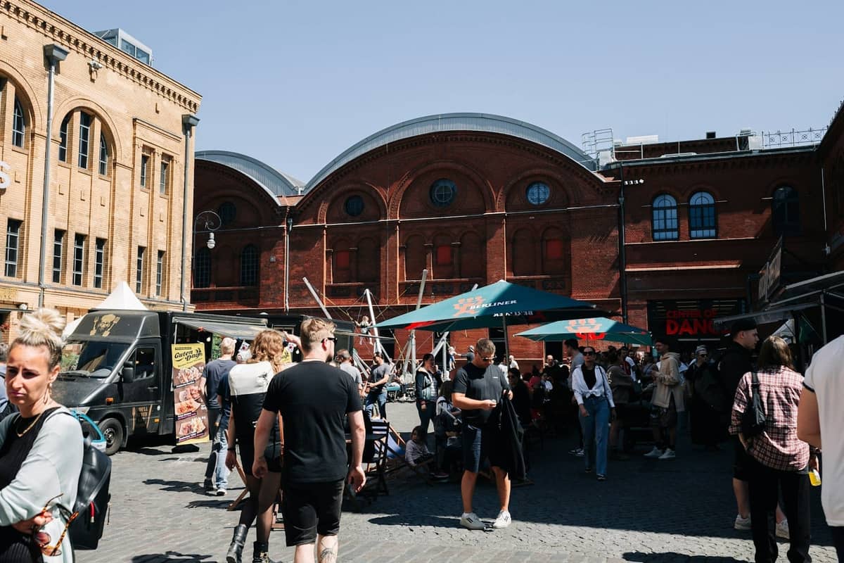 Concurrido mercado de comida al aire libre en Berlín con personas explorando puestos, rodeado de históricos edificios de ladrillo rojo y camiones de comida callejera.
