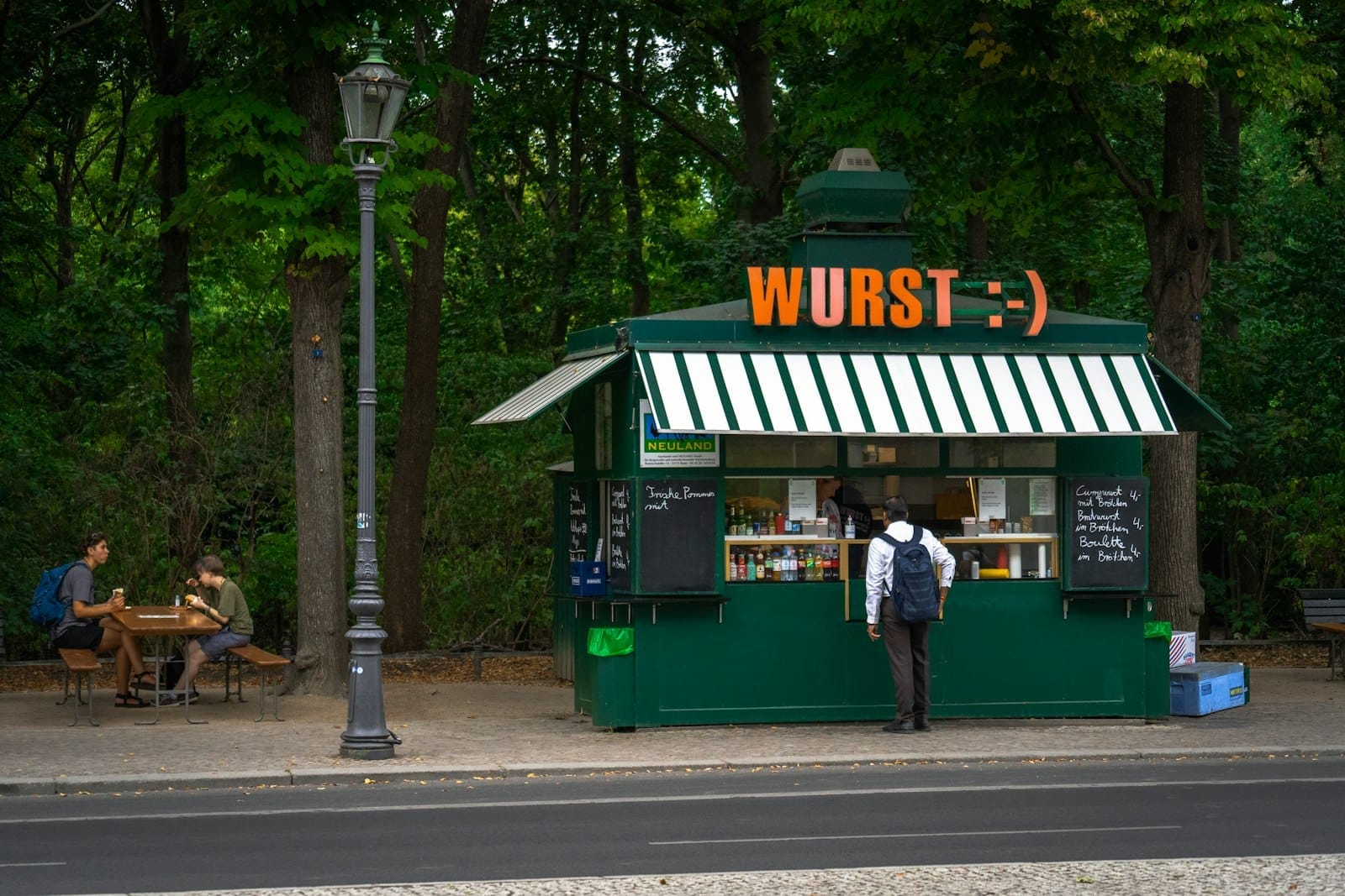 Uma barraca verde de comida de rua em Berlim com a placa WURST, com pessoas fazendo pedidos e outras comendo em uma mesa ao ar livre, cercadas por árvores.
