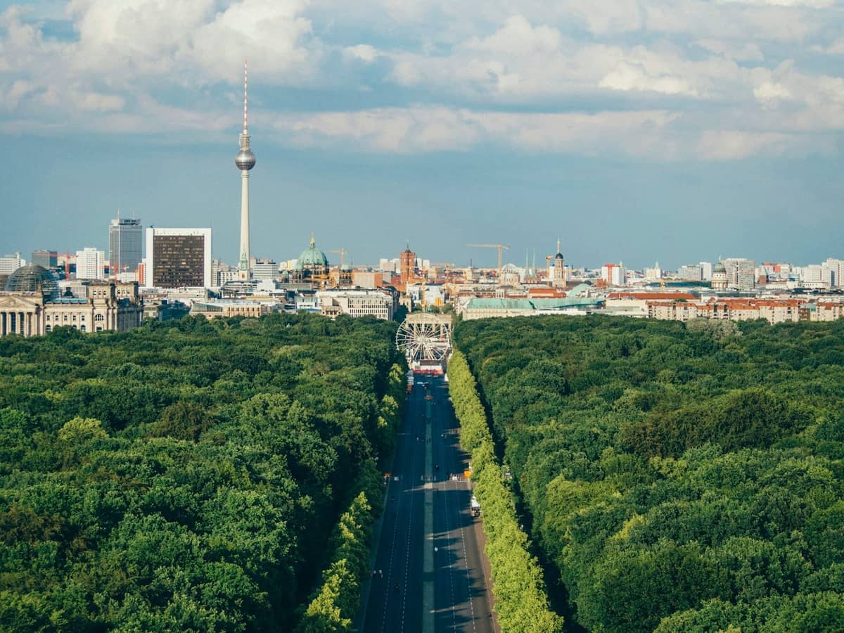 Pemandangan udara di atas taman Tiergarten Berlin dengan pepohonan hijau yang rimbun, cakrawala kota di latar belakang, dan Menara TV yang terlihat, di bawah langit yang sebagian berawan.