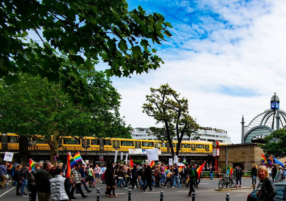 Parade jalanan yang meriah di Berlin dengan bendera pelangi, kerumunan orang, dan kereta U-Bahn Berlin berwarna kuning yang mudah dikenali di atas, pohon-pohon, serta arsitektur kota.