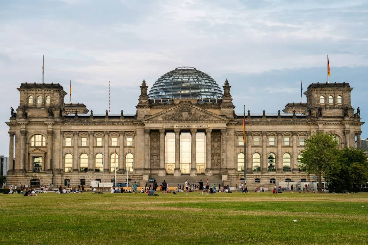 Pemandangan luas gedung Reichstag Berlin dengan kubah kacanya dan kerumunan besar di musim panas di halaman depan di bawah langit cerah.