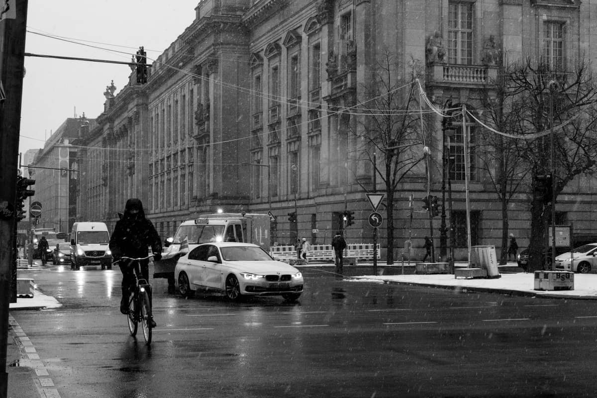 Strada di Berlino in inverno con leggera neve a terra, ciclista, automobili e grandi edifici storici sotto cieli nuvolosi.