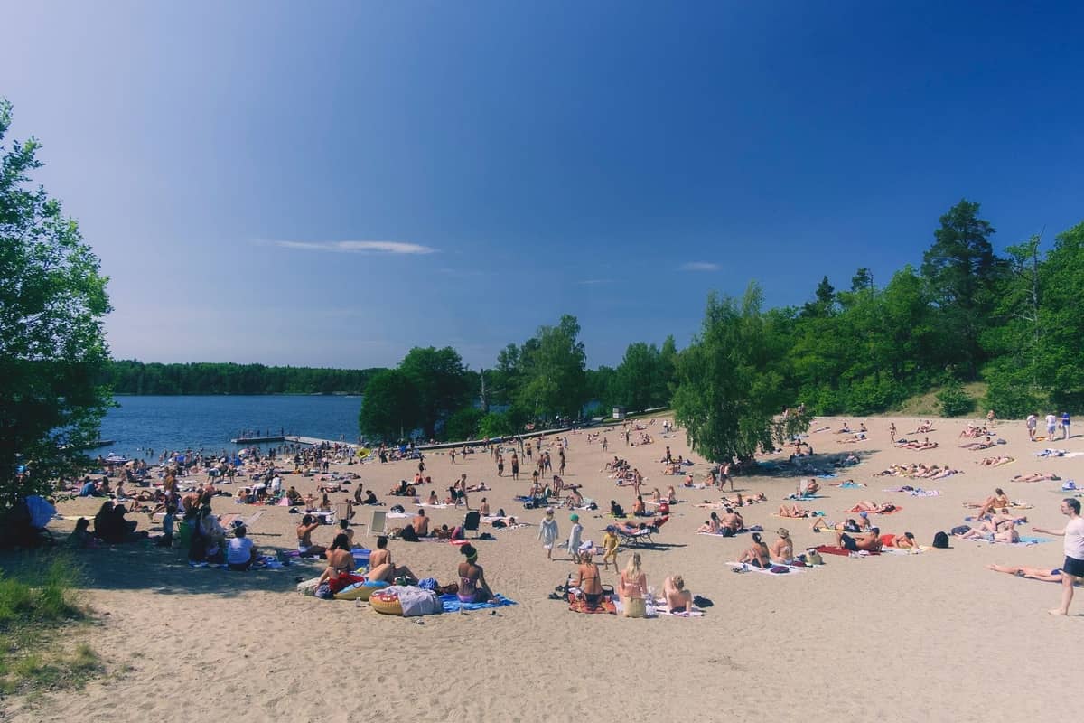 Affollata spiaggia di sabbia vicino a un lago di Berlino con molte persone che prendono il sole, nuotano e si rilassano sotto un cielo azzurro limpido.