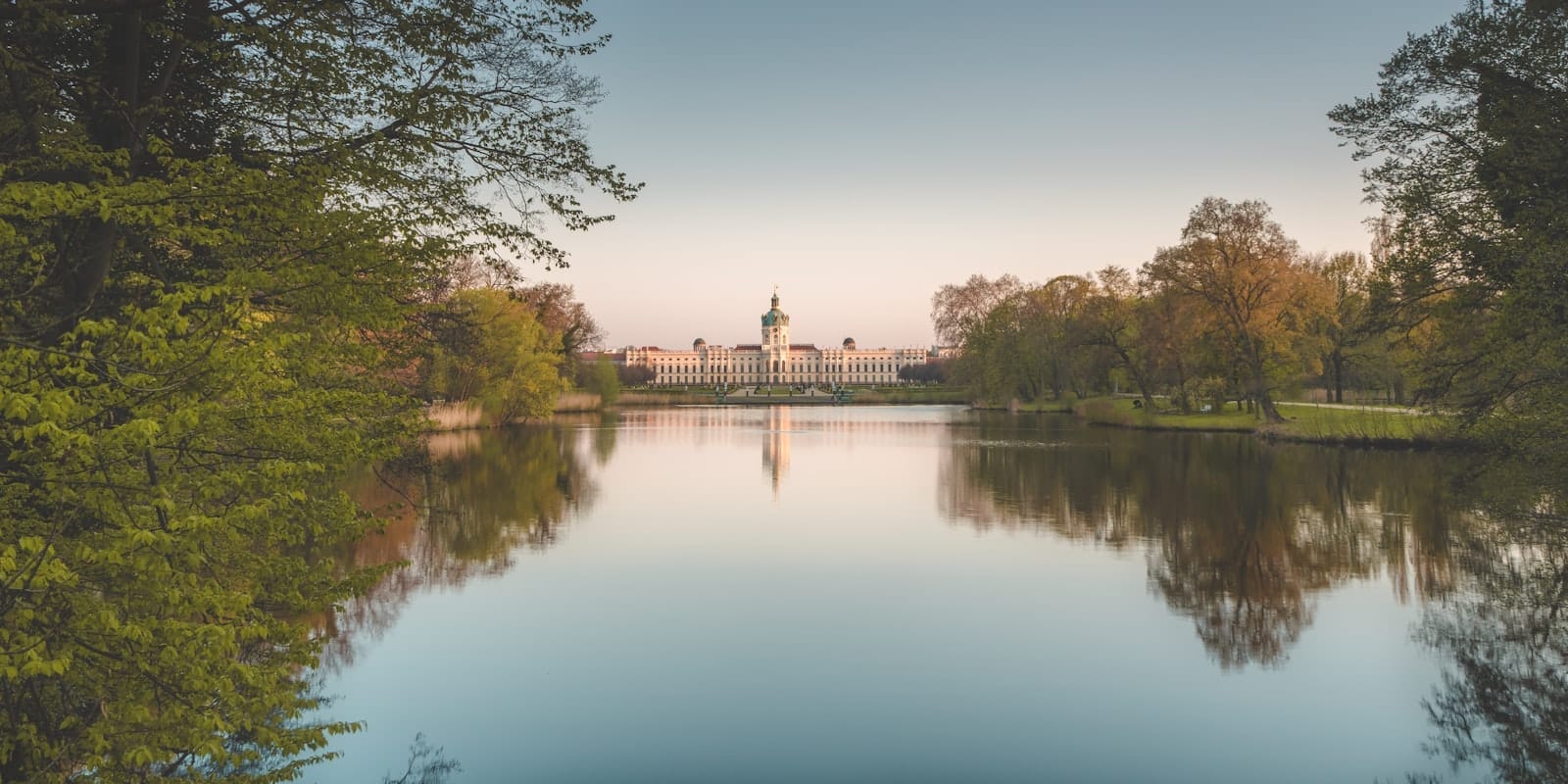 Lago tranquilo e amplo cercado por árvores verdes ao pôr do sol, com um palácio grandioso visível ao fundo, perto de Berlim.
