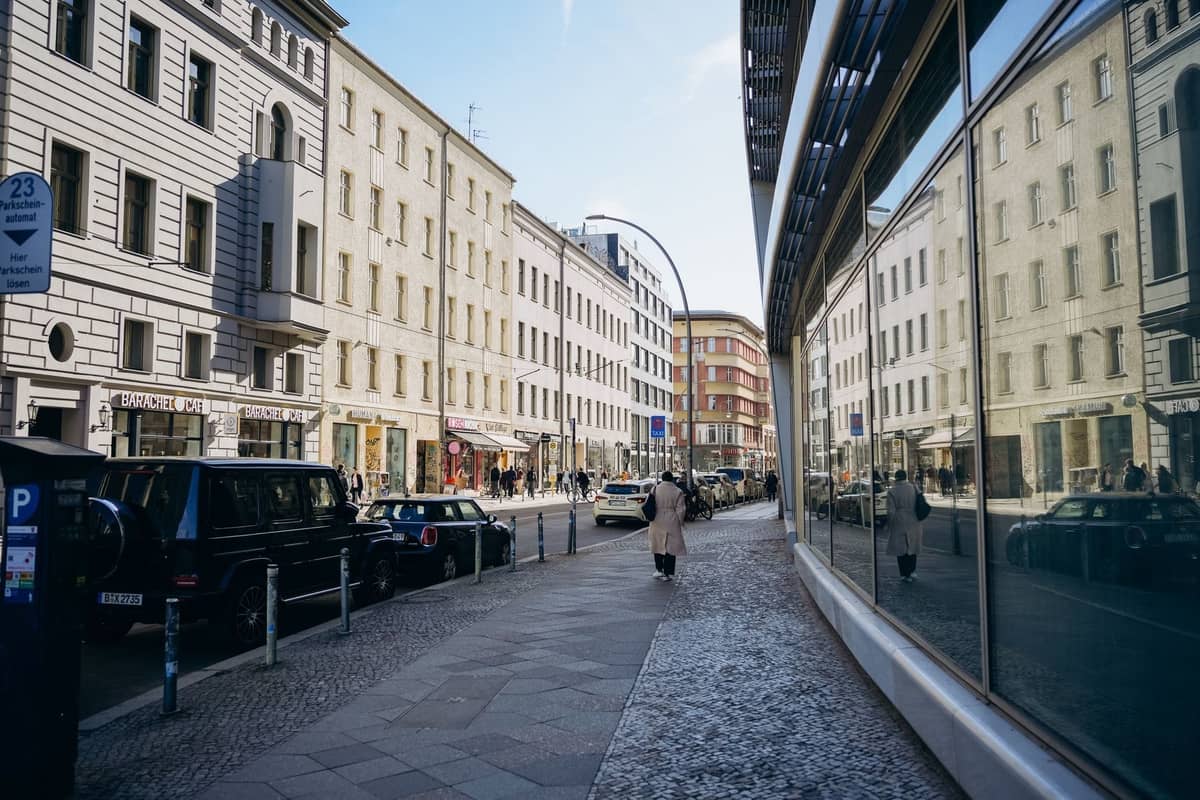 Ruhige Berliner Straße mit historischen Gebäuden, reflektierender Glaswand und Menschen, die an einem sonnigen Tag auf einem Kopfsteinpflaster-Bürgersteig spazieren.