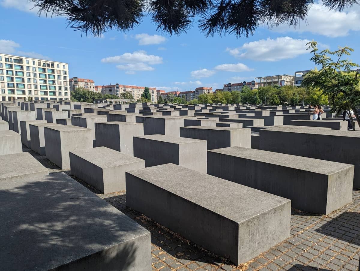 Foto des Holocaust-Mahnmals in Berlin, mit Reihen von Betonstelen, Stadtgebäuden und blauem Himmel im Hintergrund.