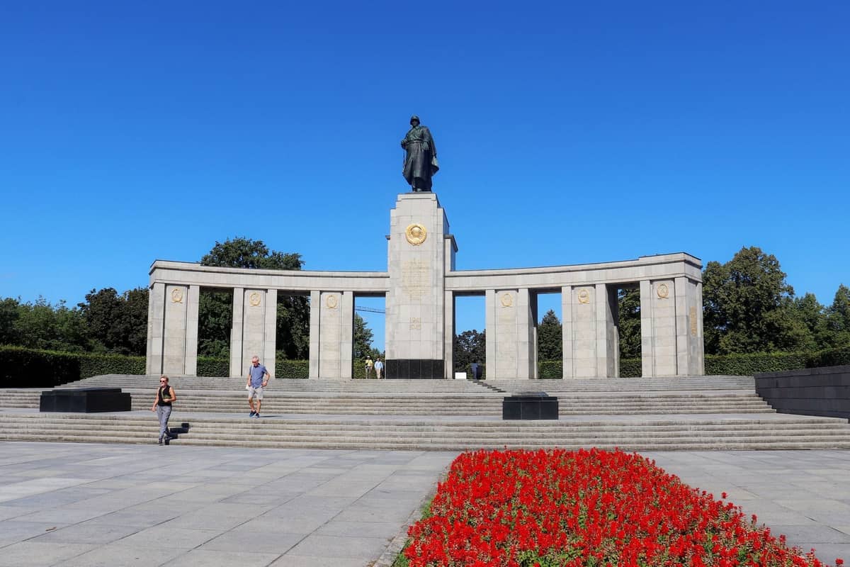 Weitläufige Ansicht des Sowjetischen Ehrenmahls in Berlin im Tiergarten, mit einer Statue, Säulen, Stufen und roten Blumenbeeten an einem sonnigen Tag.