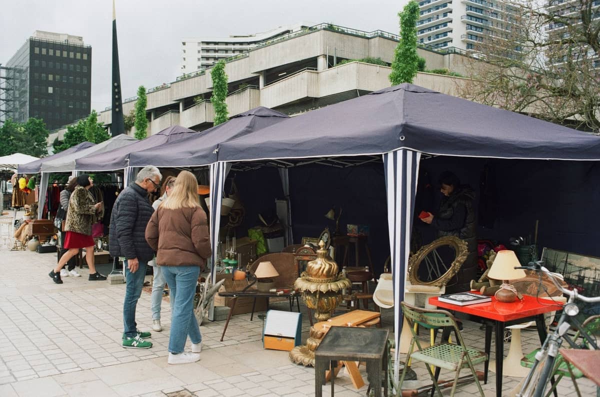 Berlin'de açık hava bit pazarında tezgahları gezen alışveriş yapanlar, arka planda modern binalar, yaz sokak yaşamı ve mevsimsel etkinlikler için tipik bir görünüm.