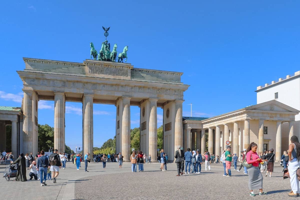 Ruchliwy plac w Berlinie z ludźmi spacerującymi i gromadzącymi się przed słynną Bramą Brandenburską pod bezchmurnym błękitnym niebem.