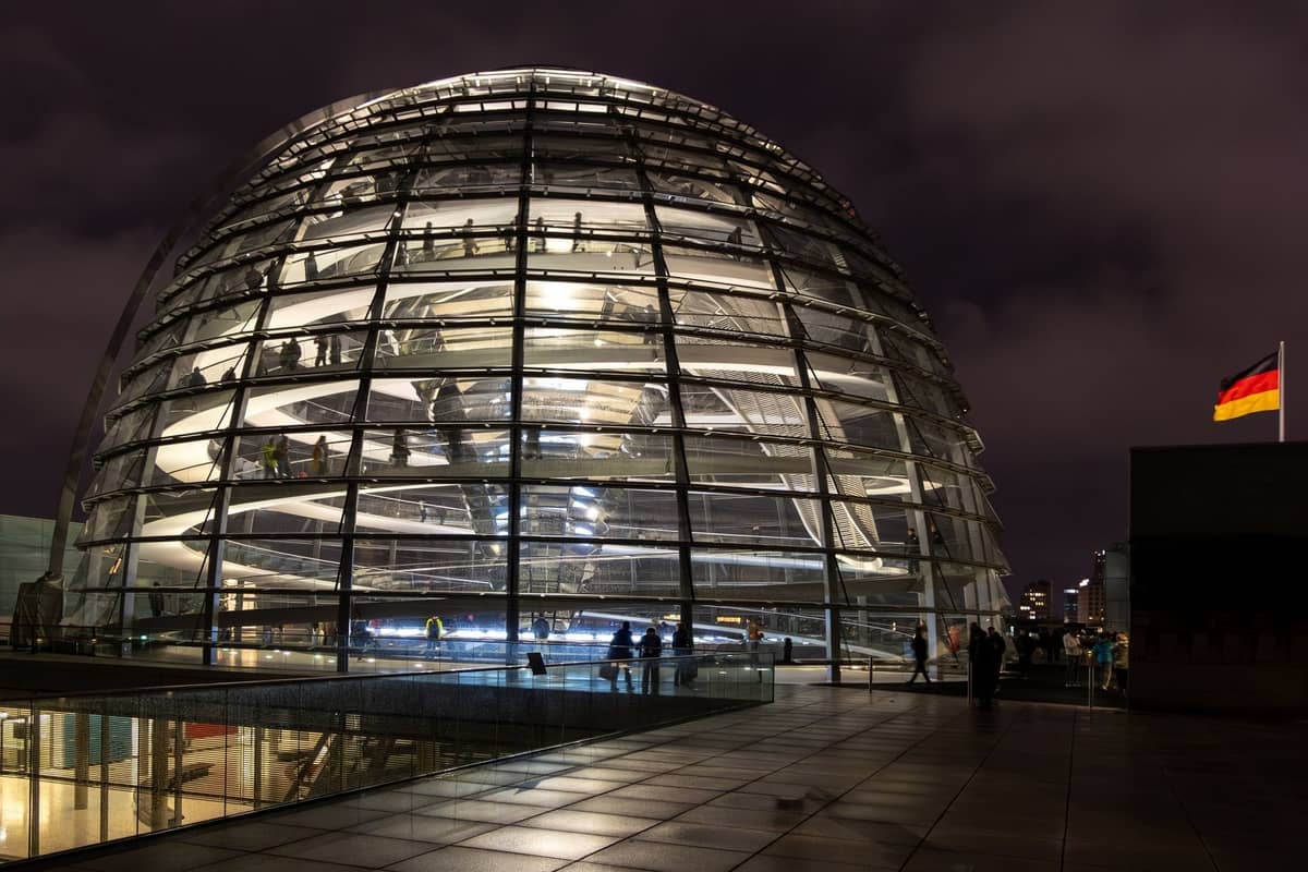 Kopuła Reichstagu oświetlona nocą z widocznymi w środku ludźmi, niemiecką flagą i zachmurzonym niebem w tle.