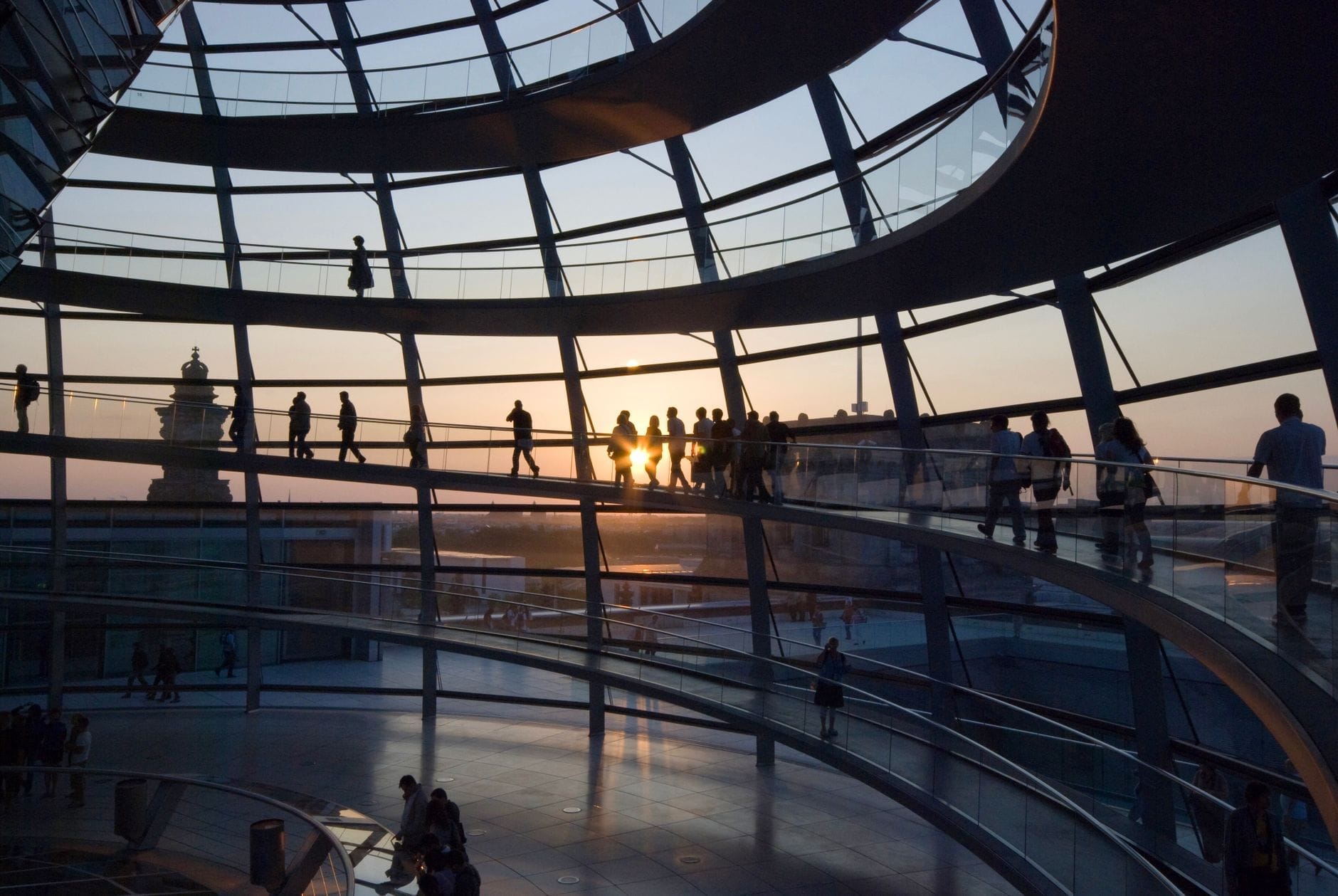 Visitantes caminhando dentro da cúpula de vidro do Reichstag ao pôr do sol, com luz natural suave iluminando as rampas curvas e o skyline de Berlim ao fundo.