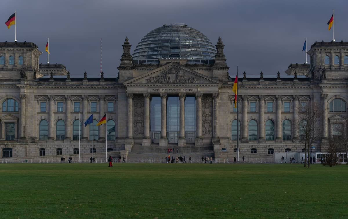 Szeroki widok na budynek Reichstagu w Berlinie z jego szklaną kopułą, niemieckimi flagami i ludźmi przed nim na tle zachmurzonego nieba.