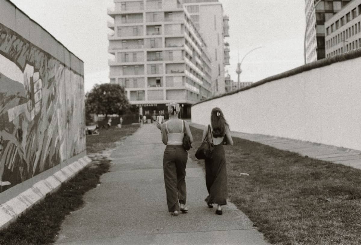 Zwei Personen, die entlang der East Side Gallery in Berlin spazieren, mit Wandmalereien und Stadtgebäuden im Hintergrund.