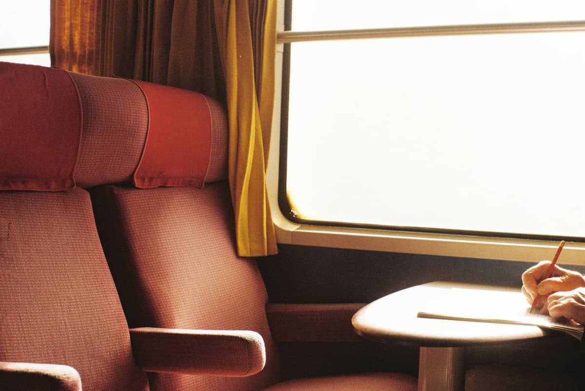 Interior de un cómodo vagón de tren de larga distancia con asientos rojos, una mesa y una persona escribiendo junto a la ventana.