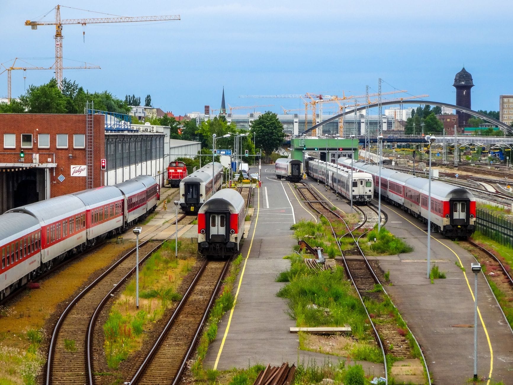 Vários trens estilo EuroCity em um movimentado pátio ferroviário em Berlim, com trilhos, plataformas e o skyline da cidade ao fundo sob um céu azul.