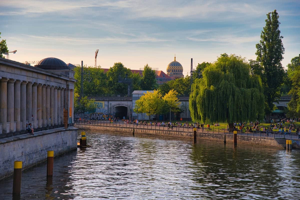 Blick auf die Uferpromenade in Berlin mit einem Säulengebäude, einem grünen Park und Menschen, die sich im Abendlicht am Wasser entspannen.