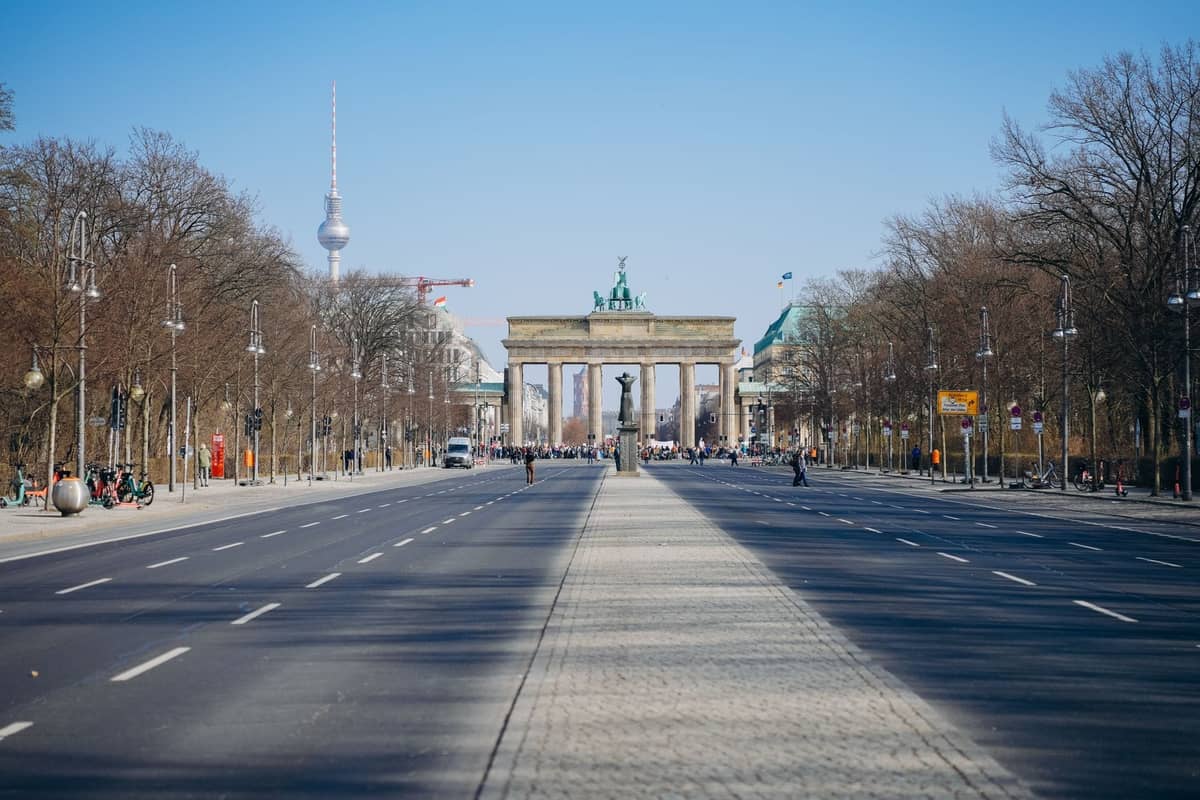 Weitwinklige Straßenansicht entlang der Unter den Linden in Richtung Brandenburger Tor, mit dem Berliner Fernsehturm im Hintergrund und Menschen in der Ferne.