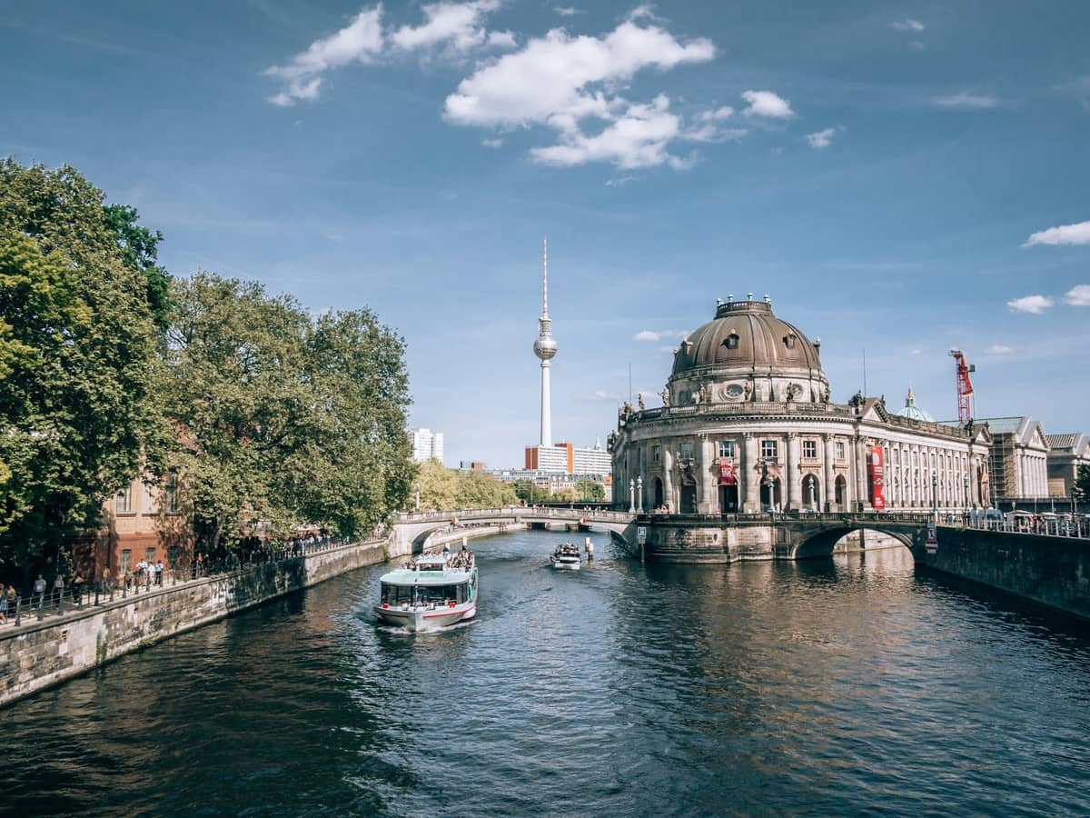 Blick auf die Berliner Museumsinsel mit dem Bode-Museum, der Spree, einem Ausflugsboot und dem Fernsehturm unter blauem Himmel