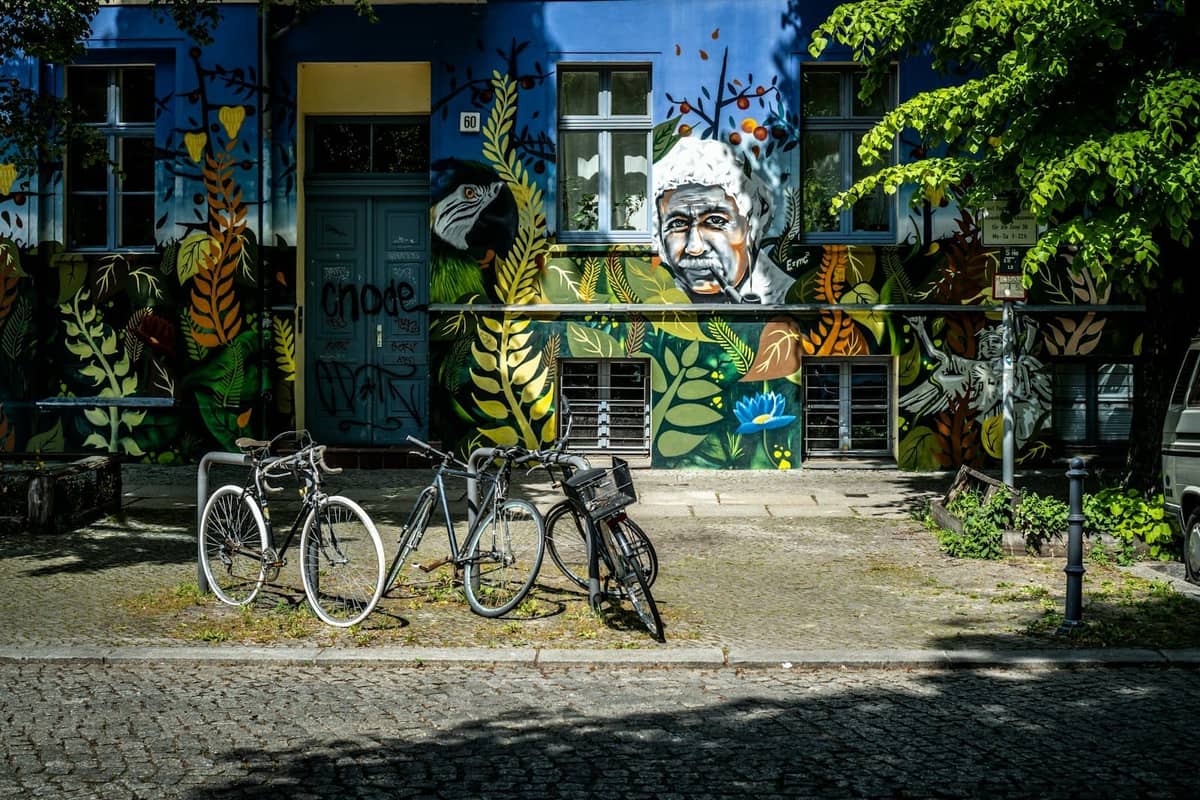 Straßenszene mit buntem Wandgemälde und Porträt an einem Gebäude in Berlin, davor geparkte Fahrräder, Bäume und Kopfsteinpflaster-Gehweg.