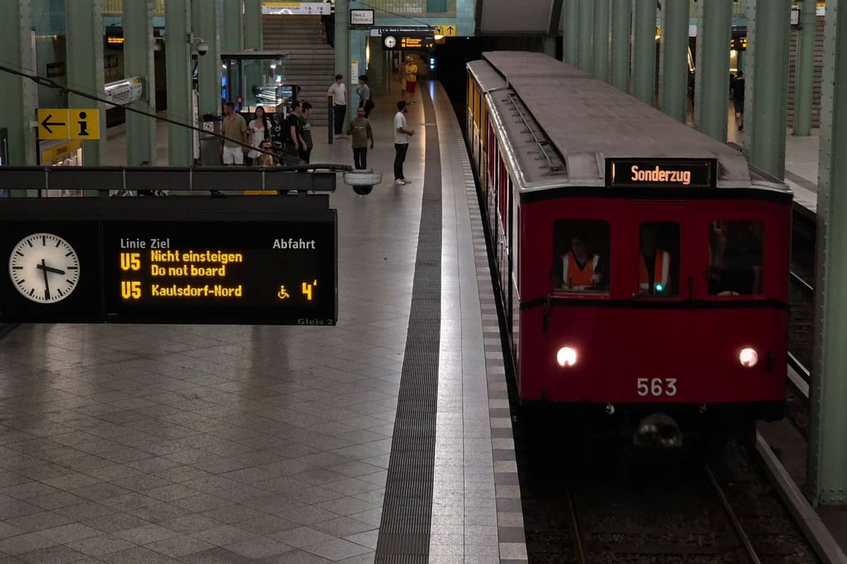Ein Berliner U-Bahnhof mit einem roten U-Bahn-Zug, digitalem Fahrplan, Uhr und wartenden Fahrgästen auf dem Bahnsteig.