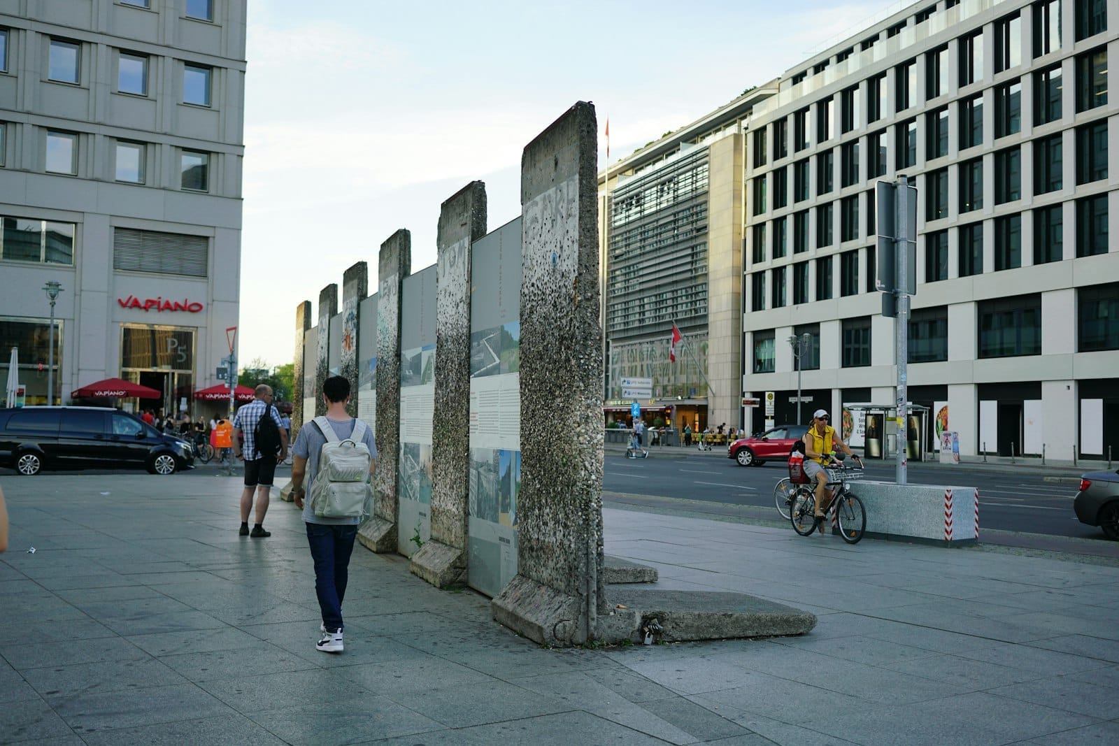 Segmentos de concreto do Muro de Berlim com grafites e murais em pé numa praça da cidade, cercados por edifícios modernos e pedestres.