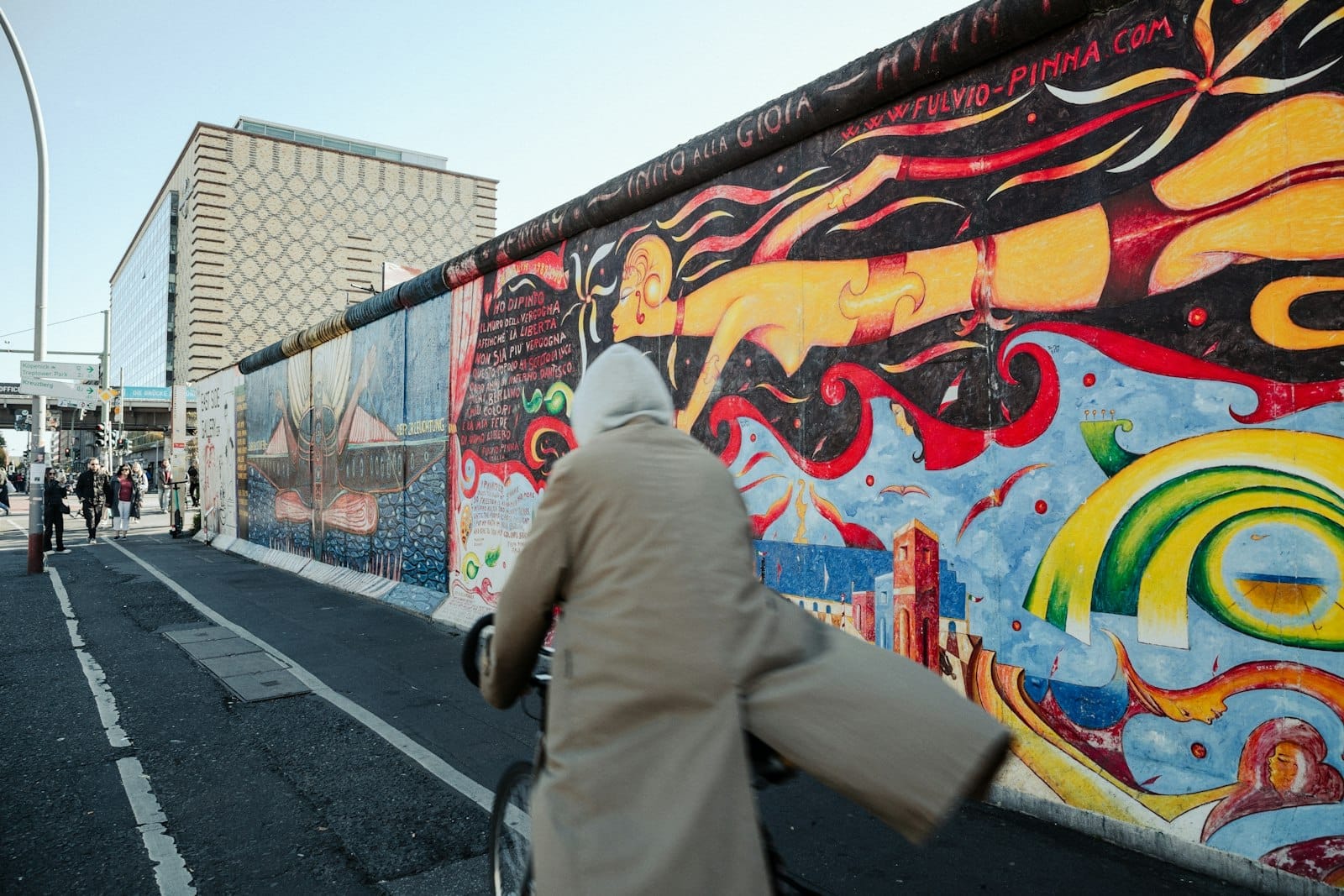 Un ciclista con un cappotto beige passa davanti al Berlin Wall Memorial riccamente dipinto, con murales colorati ed edifici moderni sullo sfondo sotto un cielo sereno.