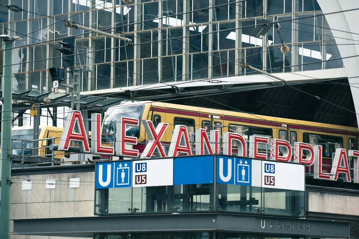 Vista de la estación de Alexanderplatz con letrero rojo, señales de transporte público y un tren pasando en Berlín.