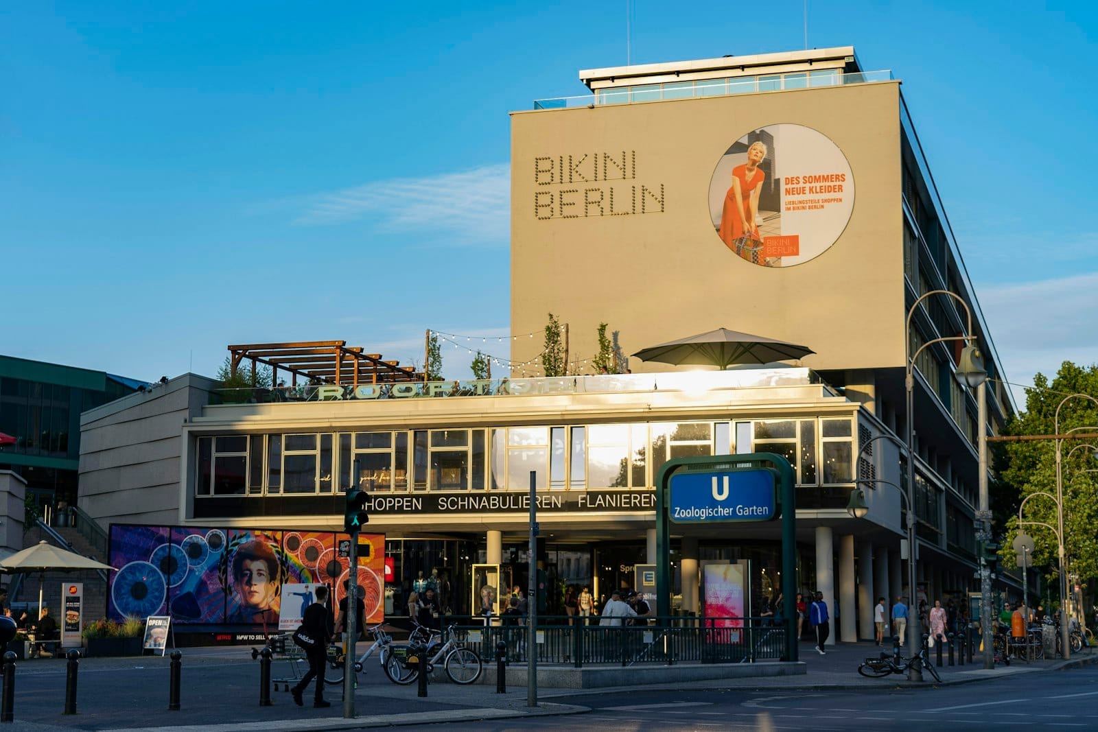 Prédio moderno com a placa 'Bikini Berlin', pessoas caminhando e pedalando, entrada do U-Bahn em primeiro plano, céu azul limpo, cena urbana vibrante em Berlim.