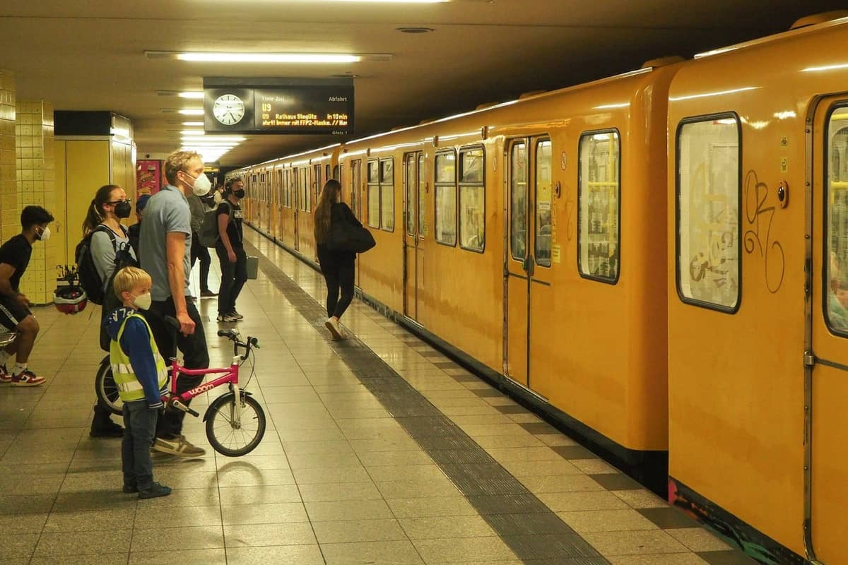 Menschen mit einem Kind und einem Fahrrad warten auf einem U-Bahn-Bahnsteig in Berlin, während ein gelber U-Bahn-Zug an der Station hält.