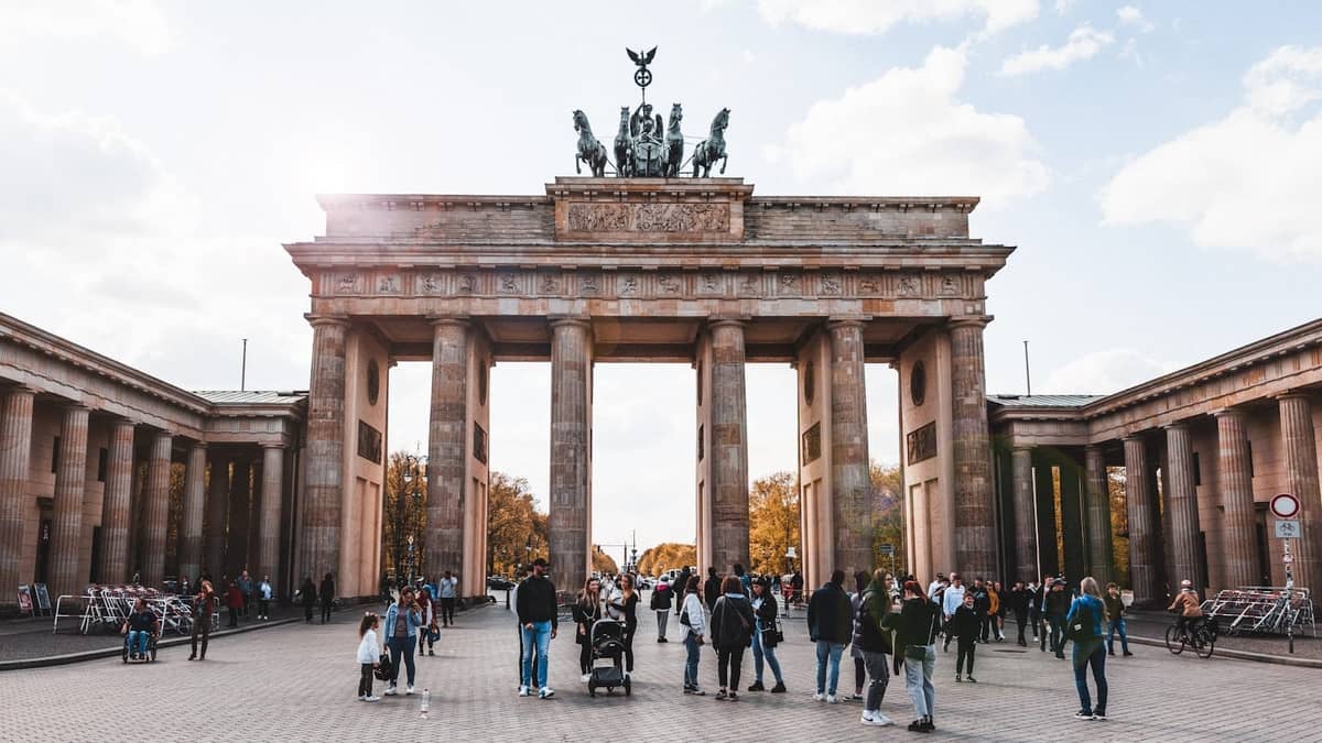 Menschen, darunter Familien mit Kinderwagen, die an einem sonnigen Tag vor dem Brandenburger Tor in Berlin spazieren und sich versammeln.