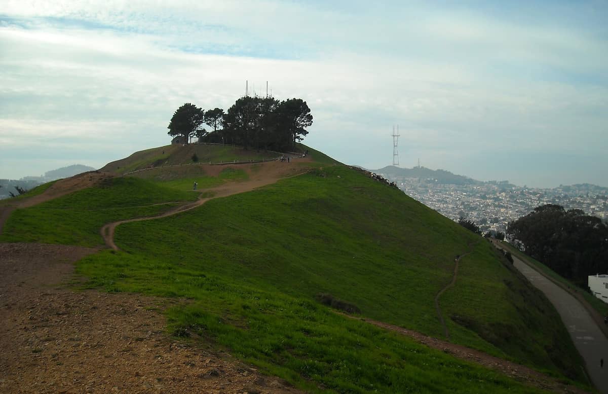 Cresta de la cima del Bernal Heights Park con personas caminando por senderos de tierra, pasto verde, árboles en lo alto y la Torre Sutro junto al paisaje urbano al fondo.