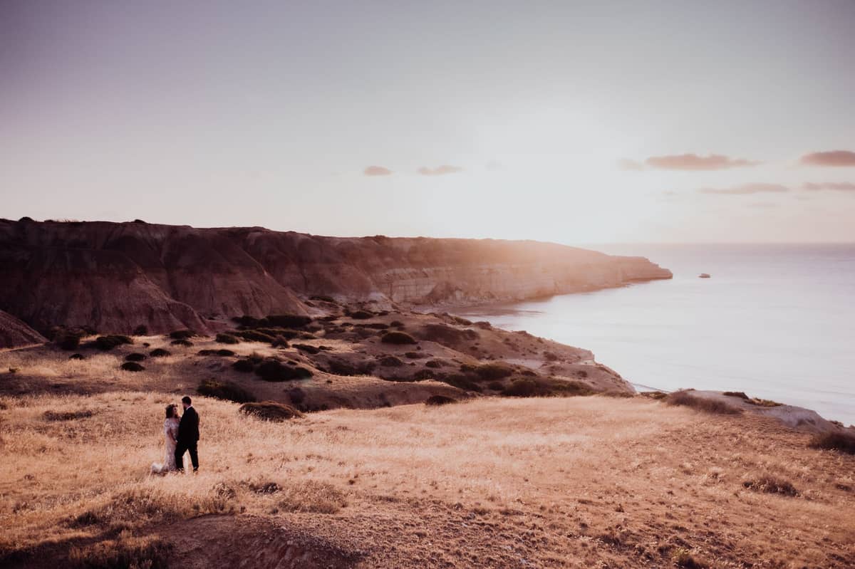 Dramatic ochre cliffs overlooking a tranquil ocean at sunset with two people standing on dry grass, typical of Fleurieu Peninsula south of Adelaide.
