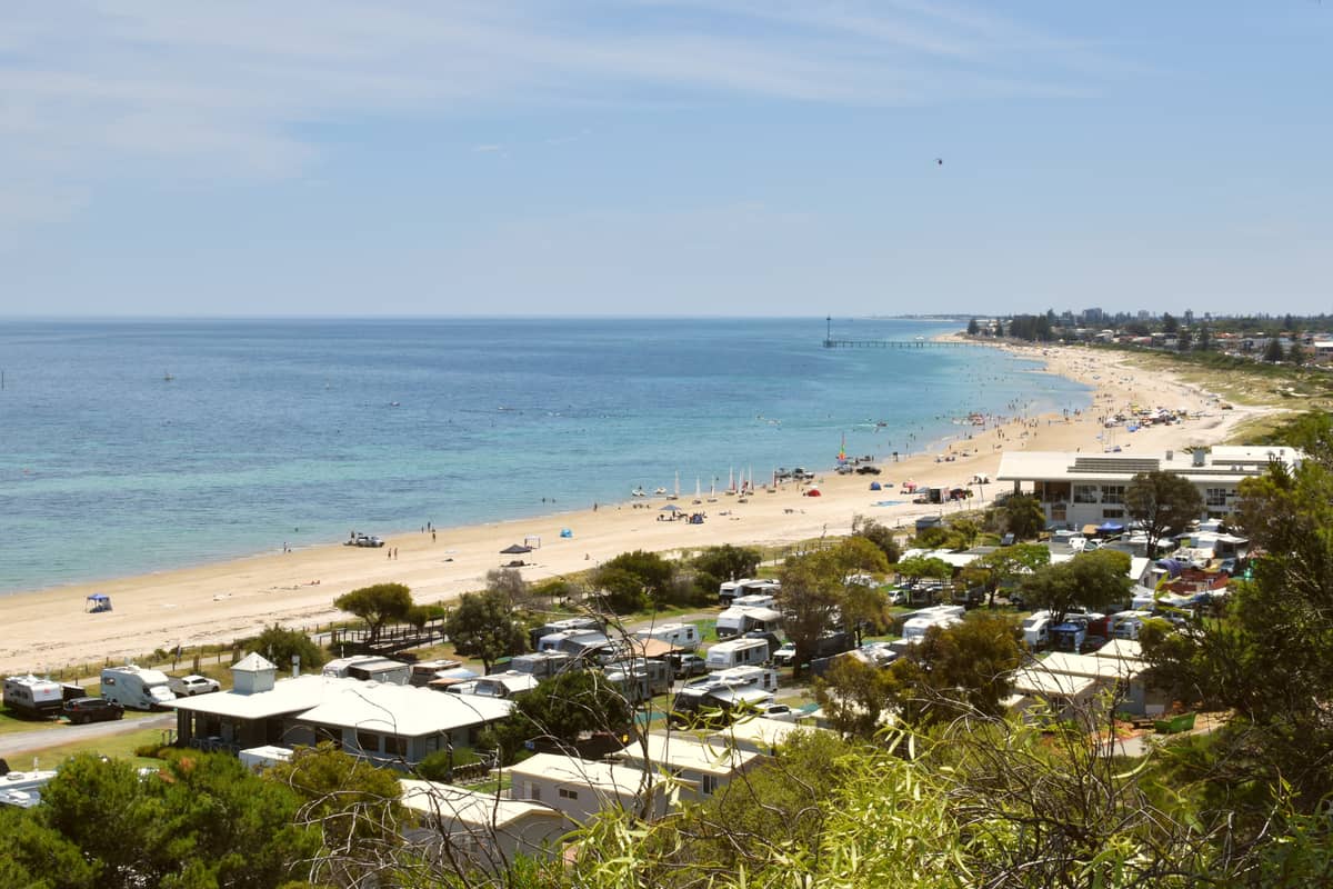 A wide view of a lively Adelaide metro beach with people, beachgoers, and distant jetty in the background.